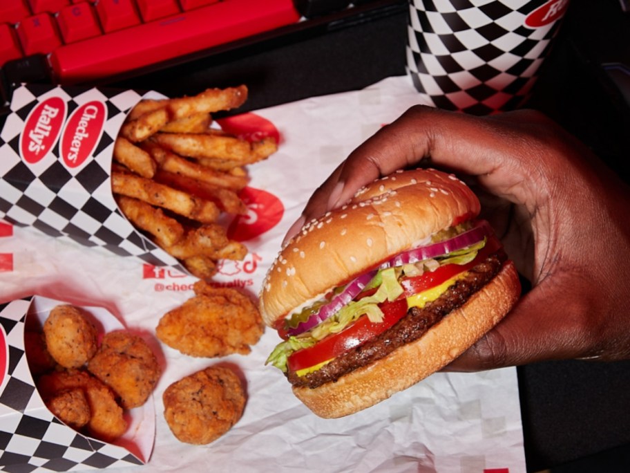 holding a Burger in front of fries, chicken nuggets, and a fountain drink from Checkers & Rally's