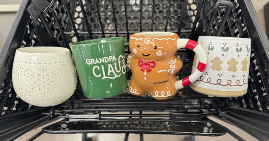 a black Kohl's shopping cart with 4 Christmas coffee mugs in the top section, a white snowflake design, a green mug with 