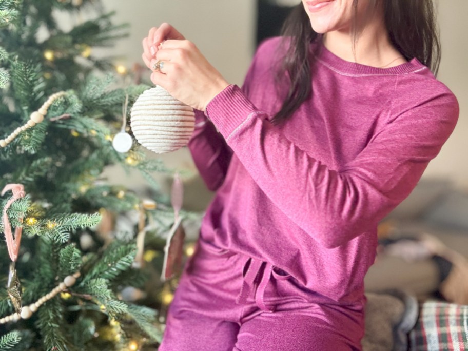 smiling woman wearing cute joyspun pjs standing next to a christmas tree and hanging an ornament on the tree