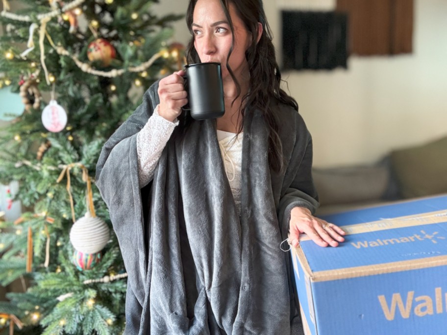 Woman sipping from warming coffee mug wearing micro plush heated throw blanket in front of Christmas tree