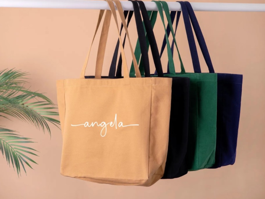 personalized canvas tote bags hanging on a rod, bags are yellow, blue, black, and green and have a name on the front