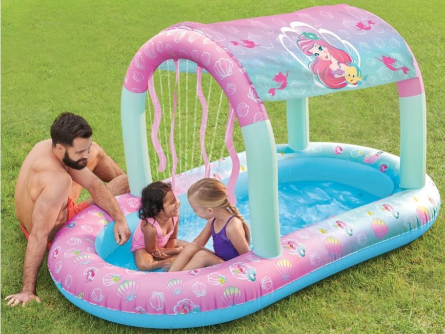 2 little girls playing in a Swimways Ariel inflatable pool with a man sitting next to the pool