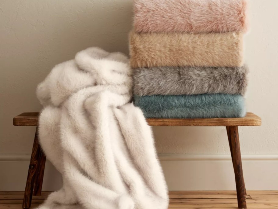 a cream faux fur blanket draped and falling off a bench, more of the same blanket in other colors stacked on the bench next to it