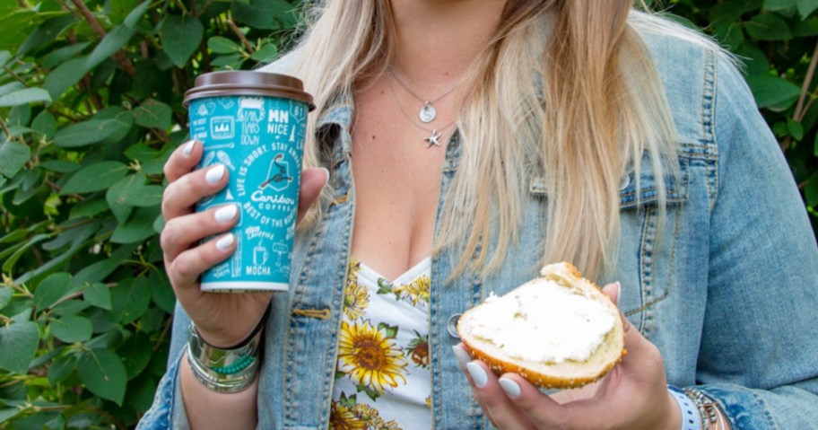 woman holding cup of coffee and half of bagel with cream cheese on it