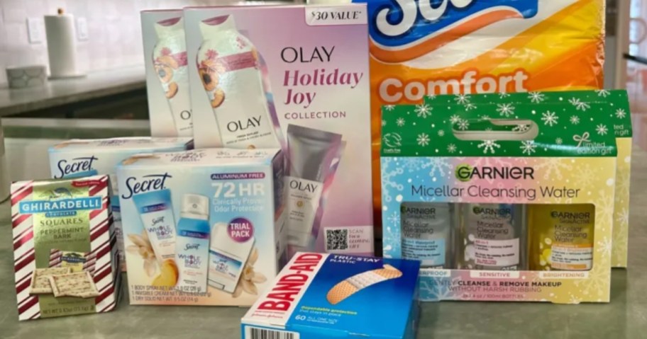 Secret, Olay, and Garnier skincare gift sets in the boxes, a box of Band-Aids, a bag of Ghirardelli chocolates and a pack of Scott toilet paper on a kitchen counter