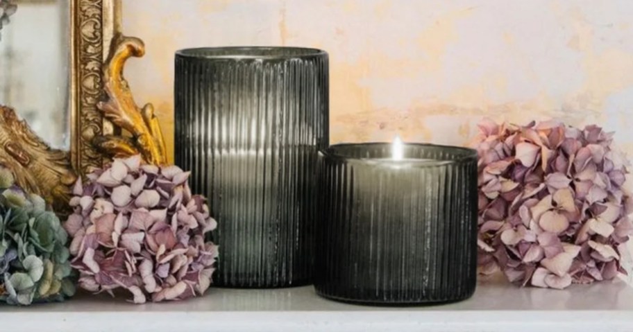 a tall and a medium size dark grey ribbed glass candle hurricane style candle holders on a mantle next to purple flowers