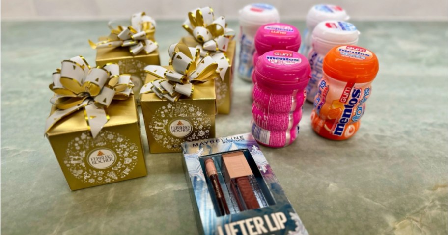 4 small gold boxes with Ferrero Rocher candy, a blue box with a Maybelline lip gloss set and 8 round containers of Mentos gum on a kitchen counter