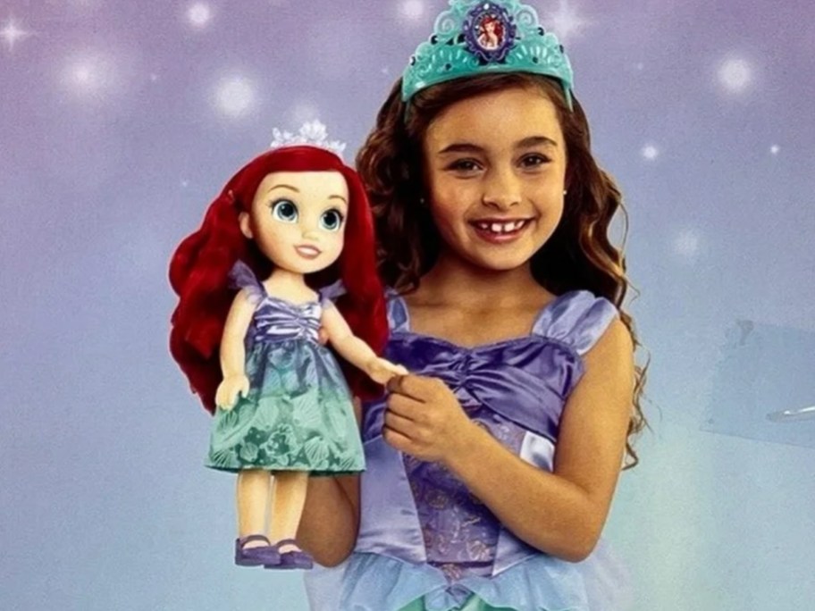 a little girl wearing a Disney Princess Ariel dress and tiara holding a matching Disney Princess Ariel doll, sparkly background behind them