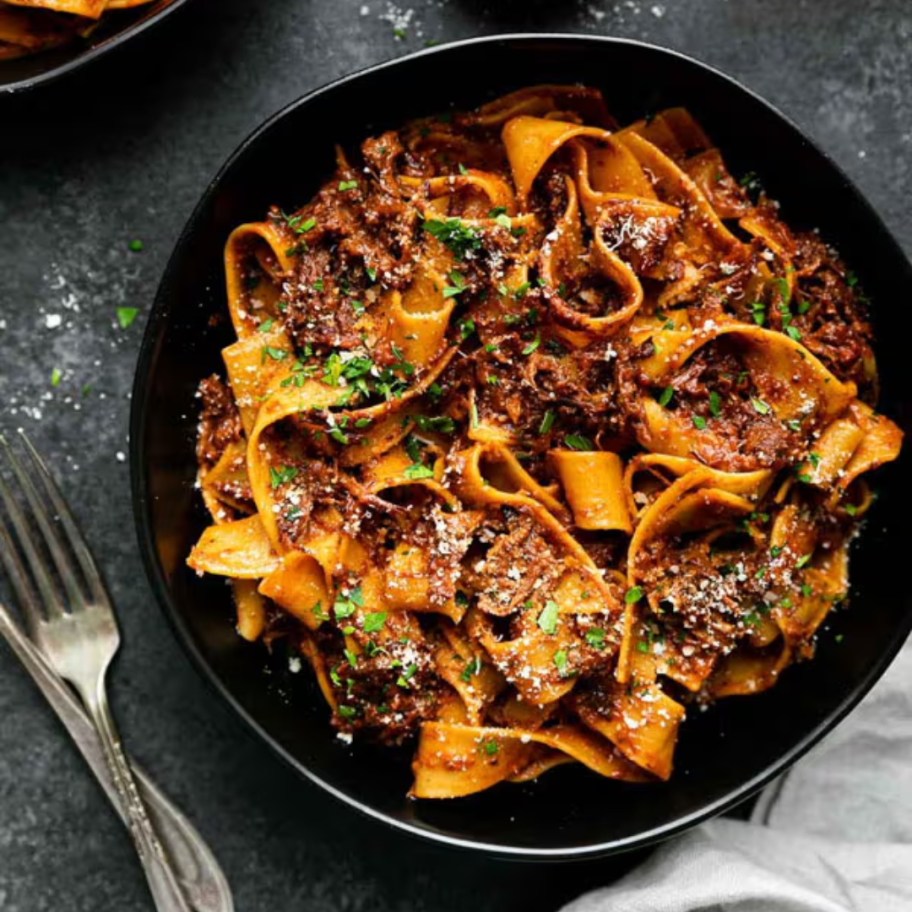 a black bowl filled with a wide pasta dish, silverware is next to it