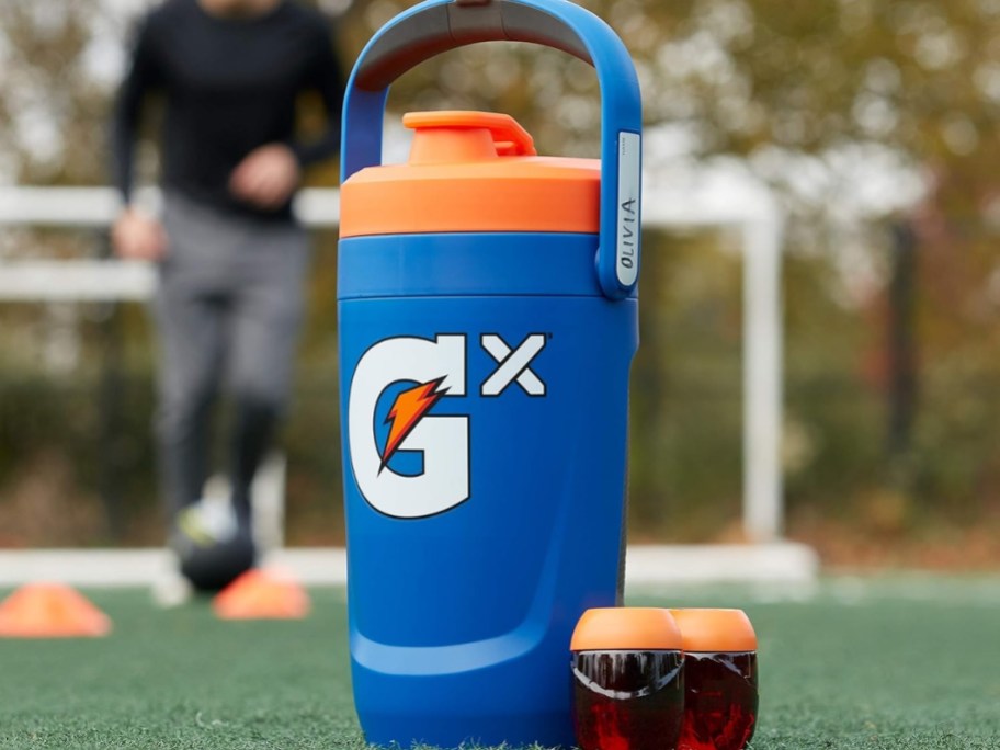 a blue Gatorade Gx large Jug with handle sitting on a soccer field, 2 Gx Pods next to it