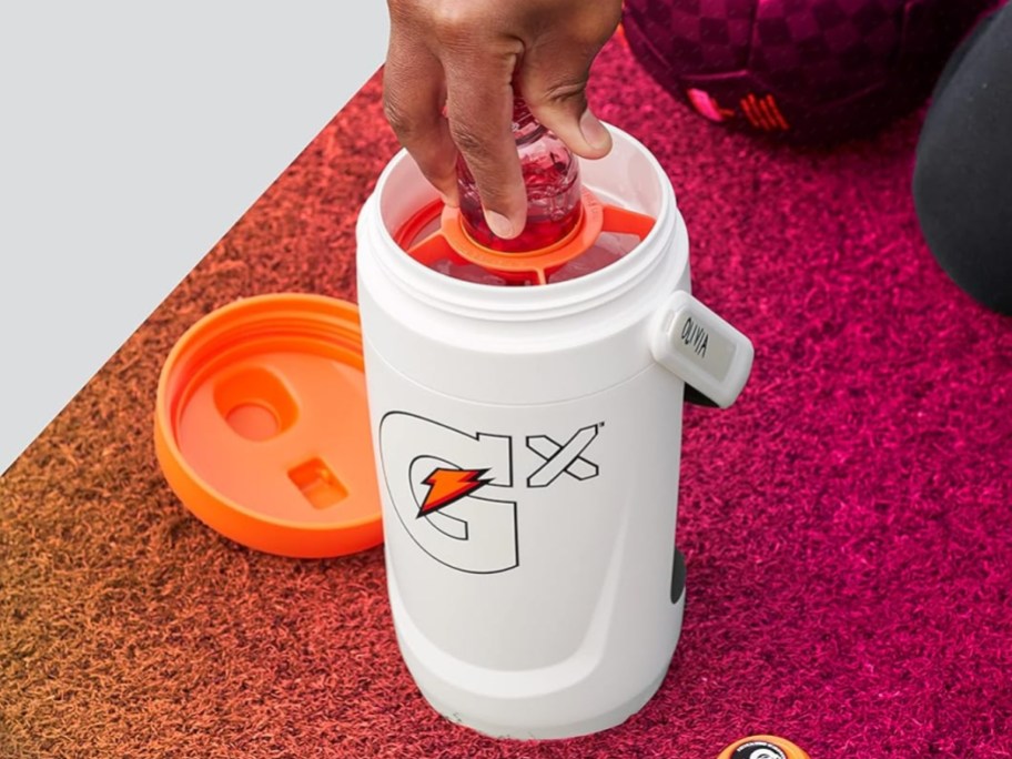 a white Gatorade Gx jug with a handle with the lid off, person is putting a Gx pod bottle with liquid in it into the jug