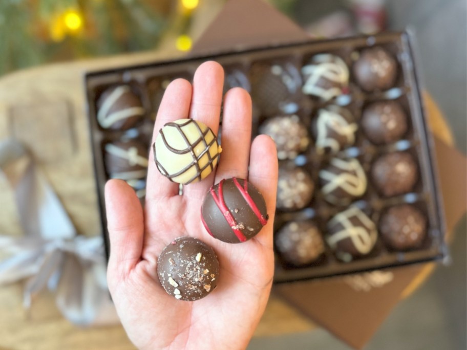 hand holding 3 different chocolate truffle candies, a box of more of them behind her hand on a table