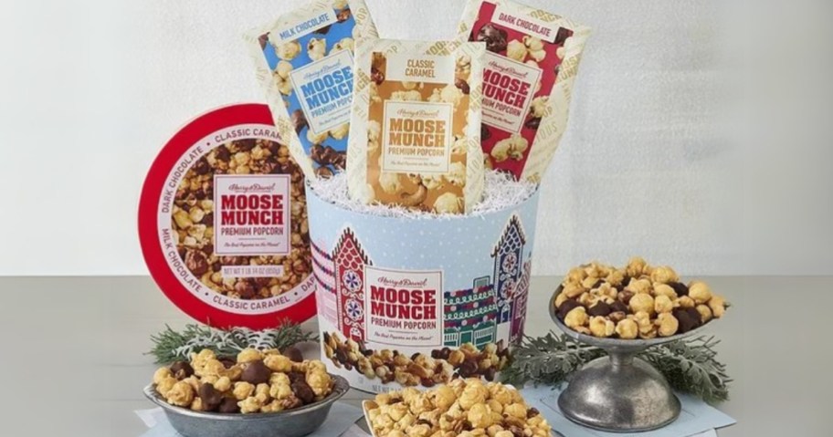 a blue and red Harry & David Moose Munch Premium Popcorn Drum with holiday print on it, open with the bags of different flavors of popcorn it comes with sticking out, there are 3 bowls in front of the drum with the different flavors of popcorn