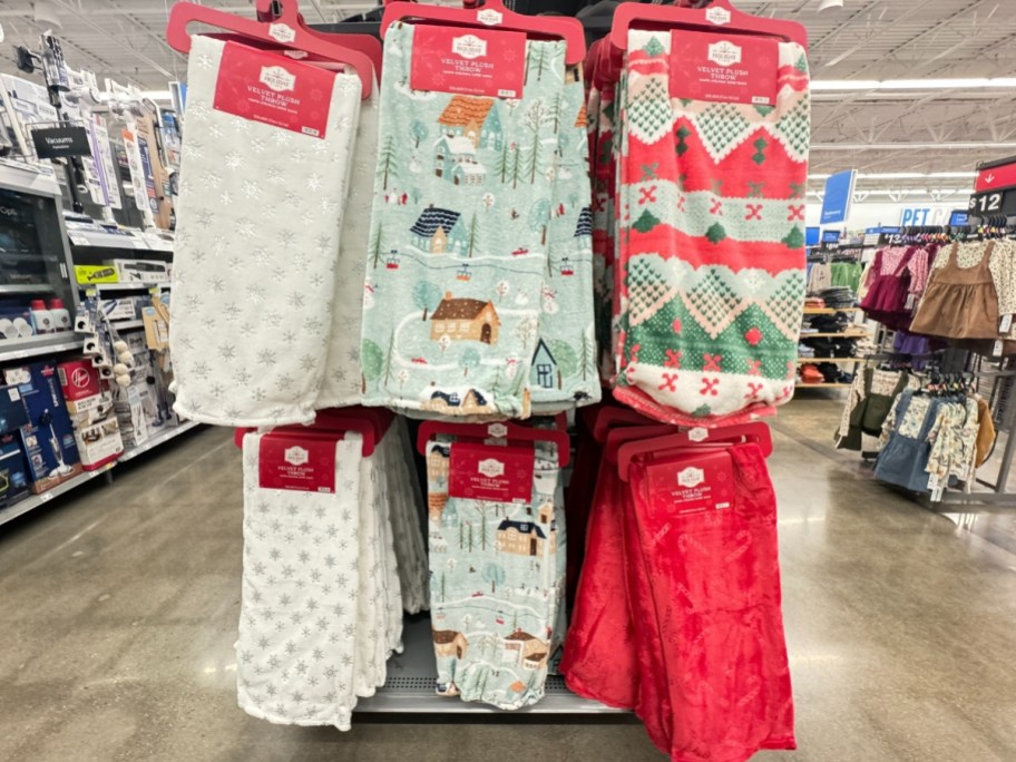 plush throw blankets on a store end cap display, white with snowflakes, light blue with winter homes and trees, one red, green, and white sweater print, and red with candy cane print