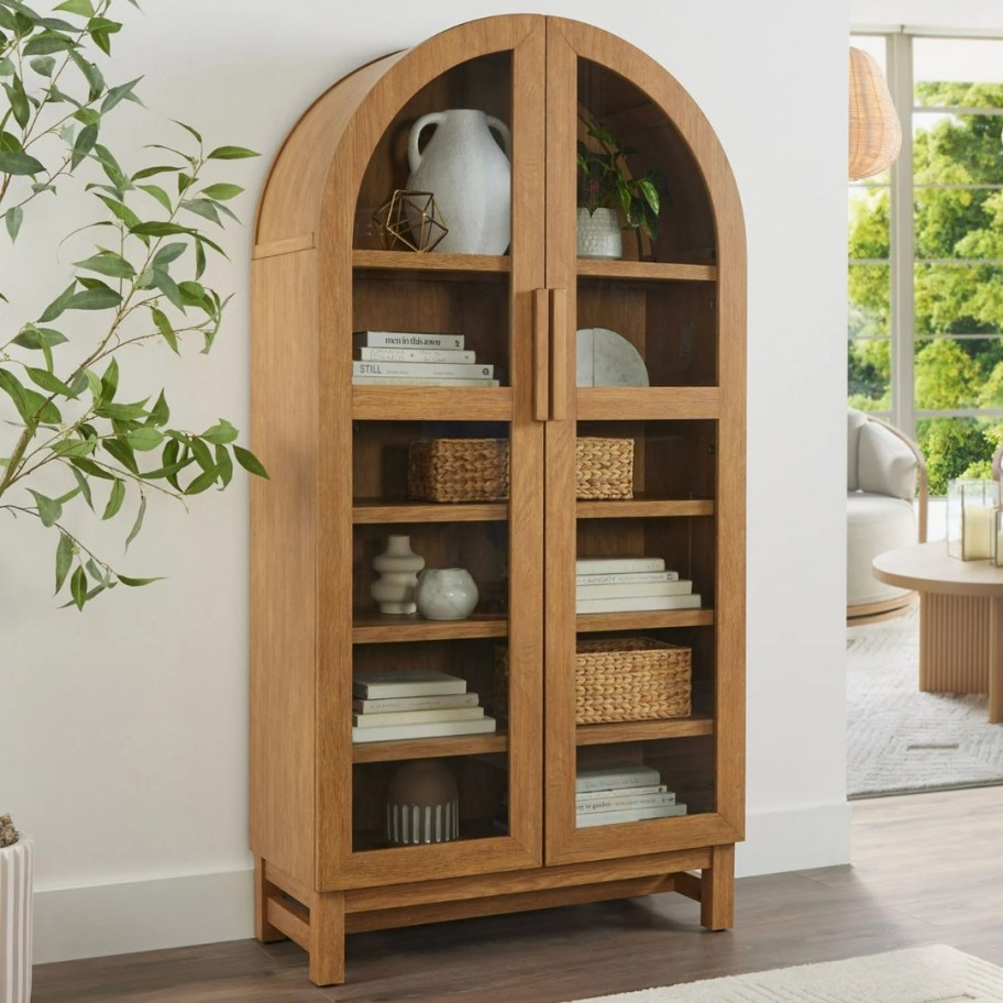 a light oak wood arched cabinet with glass doors in a hallway, living room off to the side