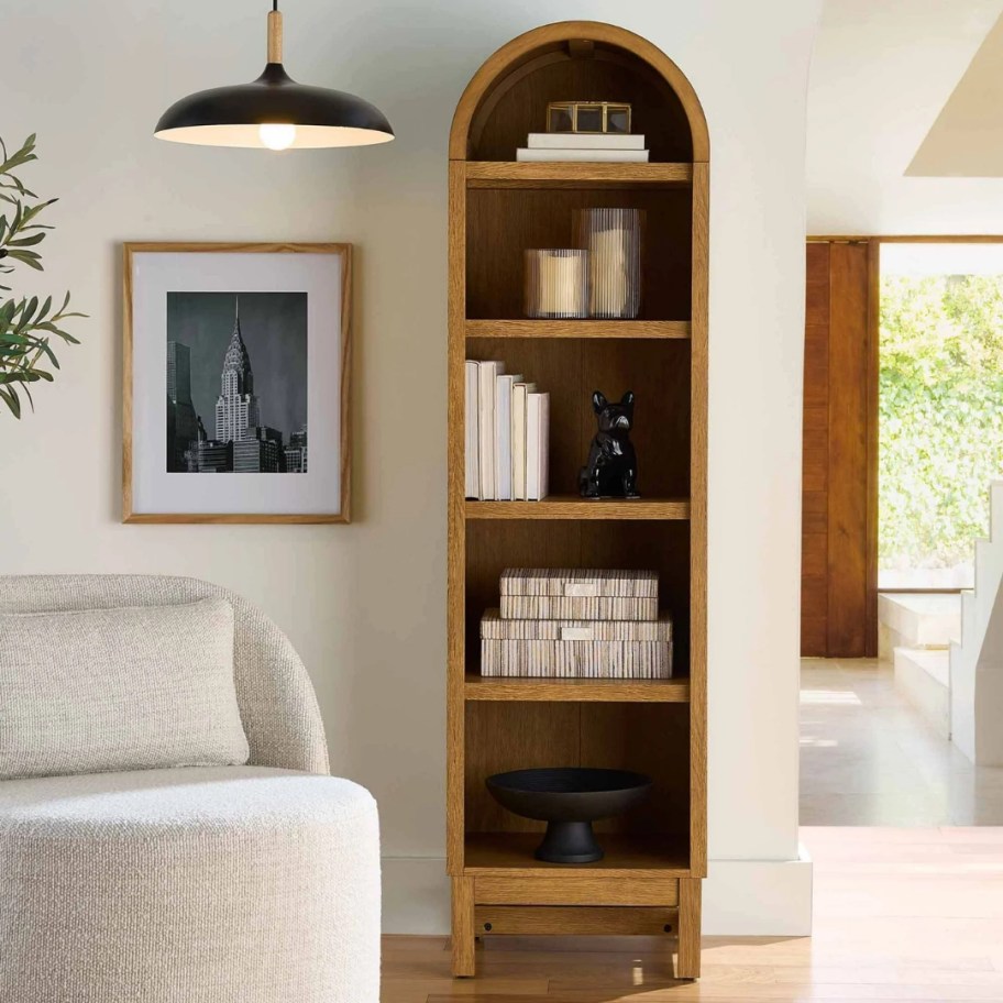 a light honey oak tone narrow arched bookcase with items on it in a living room, doorway to another room next to it