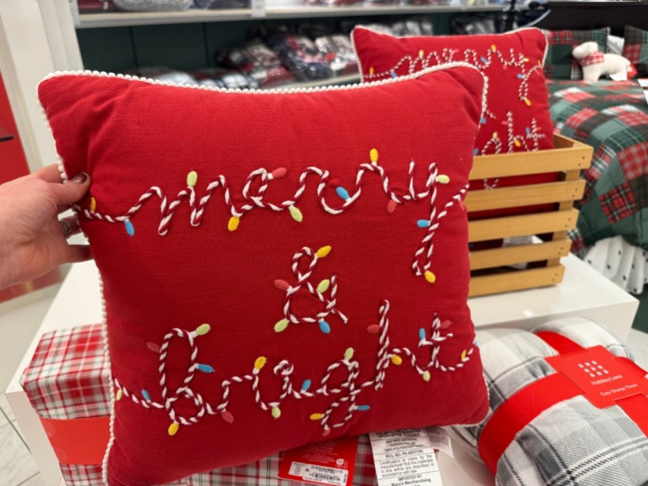 hand holding a red throw pillow that has "merry & bright" embroidered on it that looks like a string of Christmas lights