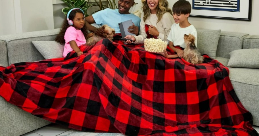 a mom, dad, little boy and little girl and 2 small tan dogs on a sofa cuddled under a red and black buffalo plaid print extra large family size blanket