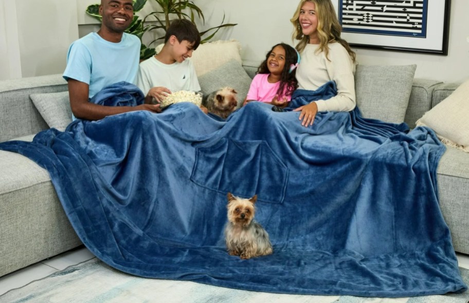 a mom, dad, little boy and little girl on a sofa cuddled under a red and black buffalo plaid print extra large family size blanket, there are 2 little tan dogs, one on the couch and on on the floor on the blanket