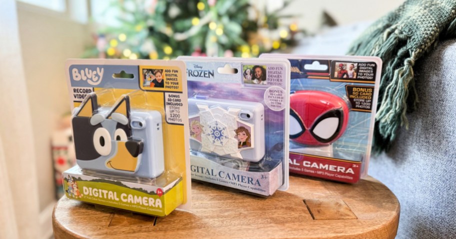 Bluey, Frozen, and Spiderman kids digital cameras on a wooden accent table in front of a Christmas tree.