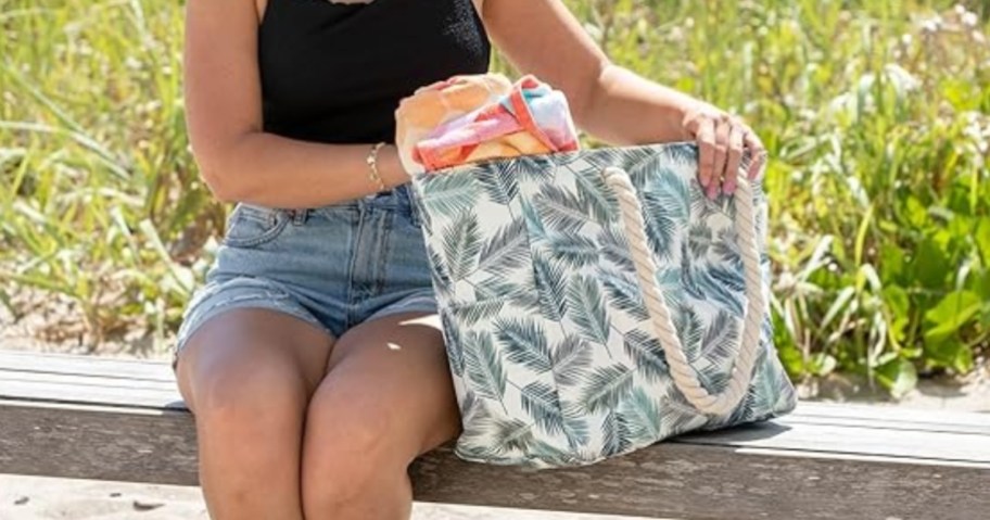 woman sitting on a bench at the beach, reaching into a large white tote bag with palm leaves print and rope handles that has beach towels and other beach items in it, there is tall sea grass and the ocean is in the background