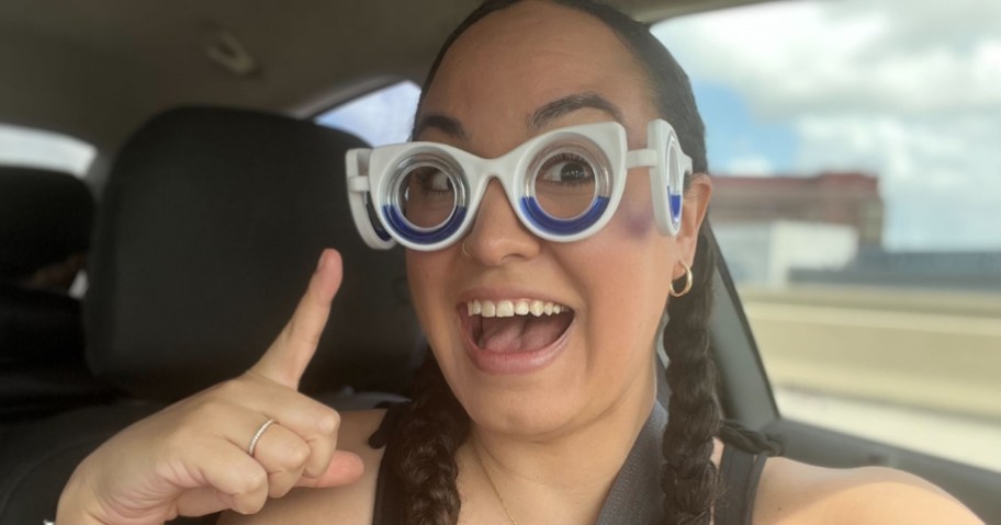 woman wearing white and blue motion sickness glasses that have lenses on her eyes and the sides of her face, she is smiling and pointing to the glasses