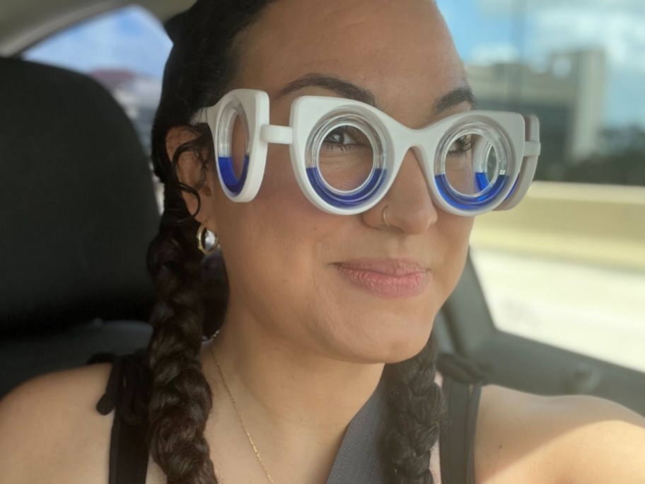 woman wearing white and blue motion sickness glasses that have lenses on her eyes and the sides of her face, she is riding in a car