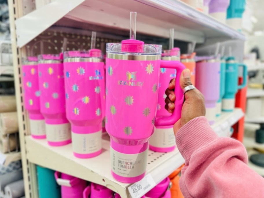 hand reaching for a bright pink Stanley tumbler that has silver stars on it, more of the same color and other pastel Stanley Tumblers on the shelf behind it