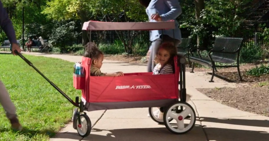 adult person pulling a red Radio Flyer wagon with a canopy with 2 kids sitting inside, another adult is walking behind it