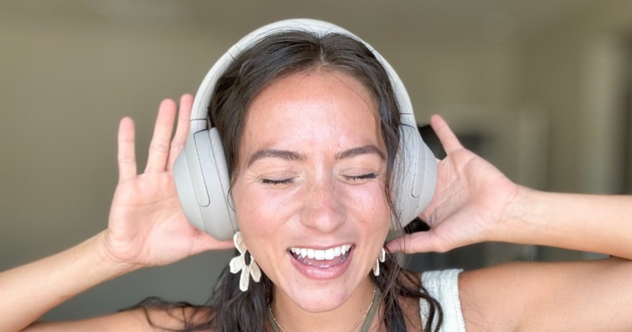 woman with brown hair wearing silver Sony headphones with her hands on them, her eyes shut and smiling