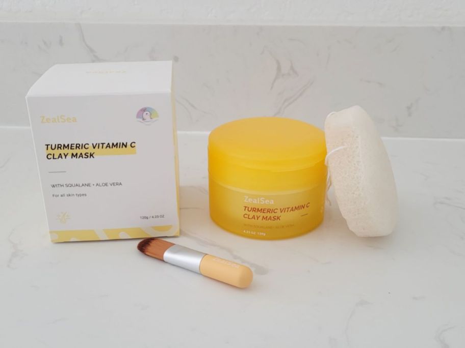 a jar of turmeric clay face mask shown with applicator brush and face puff