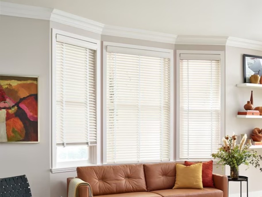 a living room with a tan couch and 3 windows behind it with white would blinds