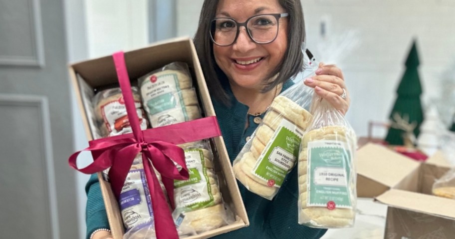 woman holding a box with a red ribbon that has bags of Wolferman's English Muffins in one hand and 2 single bags of English Muffins in her other hand, she is smiling at the camera