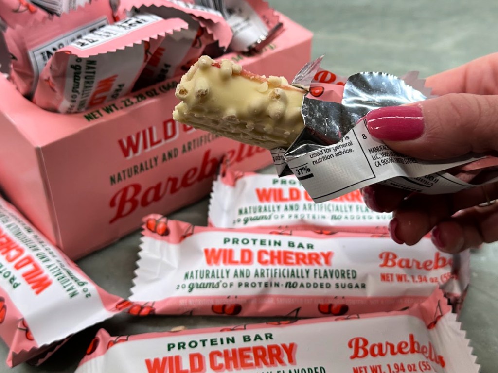 Woman holding open barbells, wild cherry protein bar in front of box