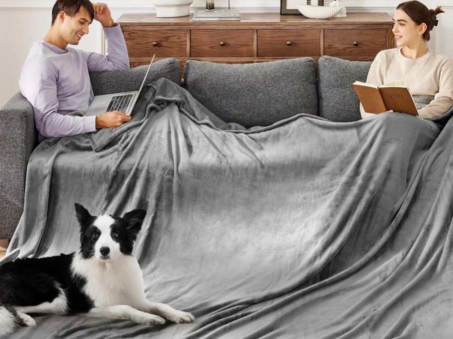 two people under gray blanket on couch with dog in front of them. 