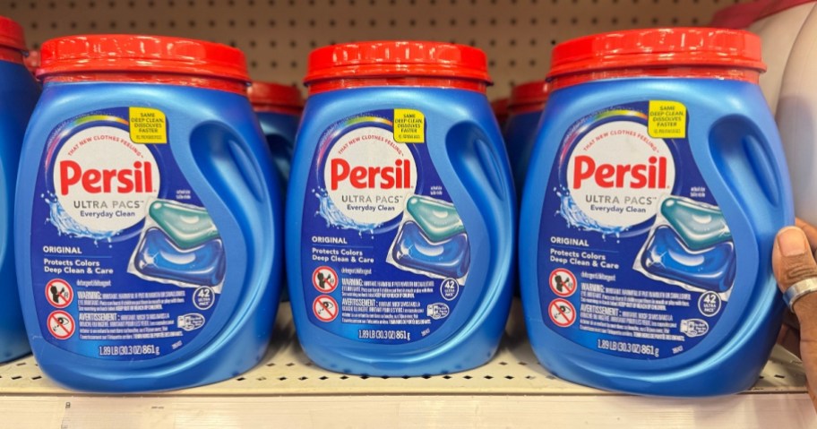 hand reaching for a blue and red container of Persil Laundry Detergent pacs, more next to it on a shelf