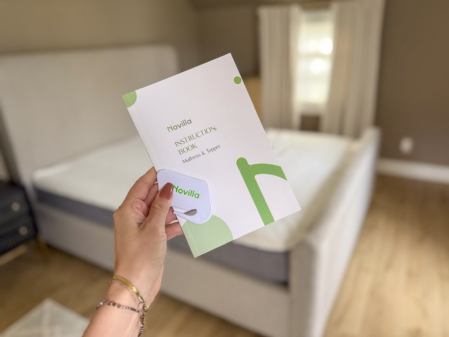 Hand holding the Novilla instruction book in front of a new novilla bliss memory foam bed