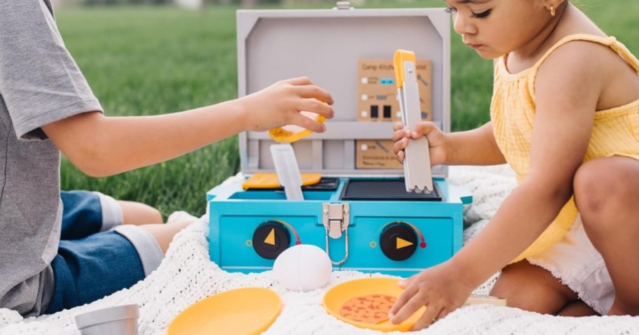 kids sitting on a blanket on the grass playing with a toy camp stove and food set