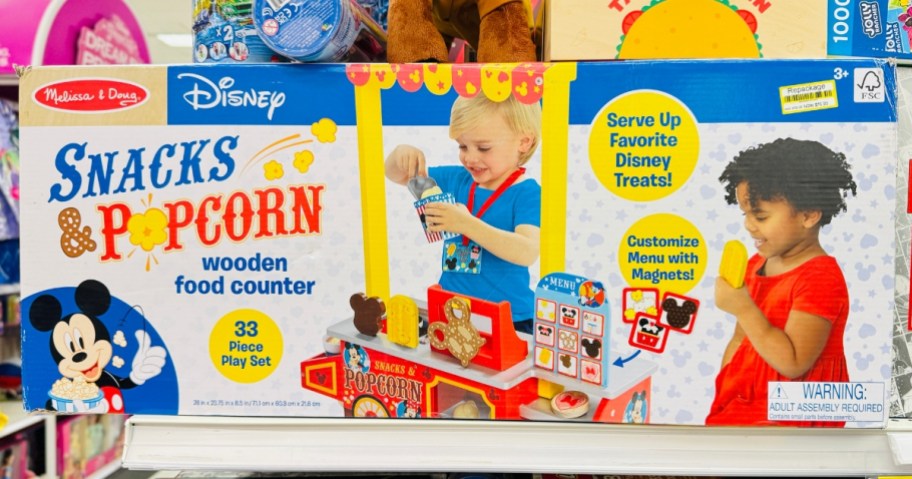 a large box on a store shelf with a Melissa & Doug Disney Snacks & Popcorn Counter Play Set in it