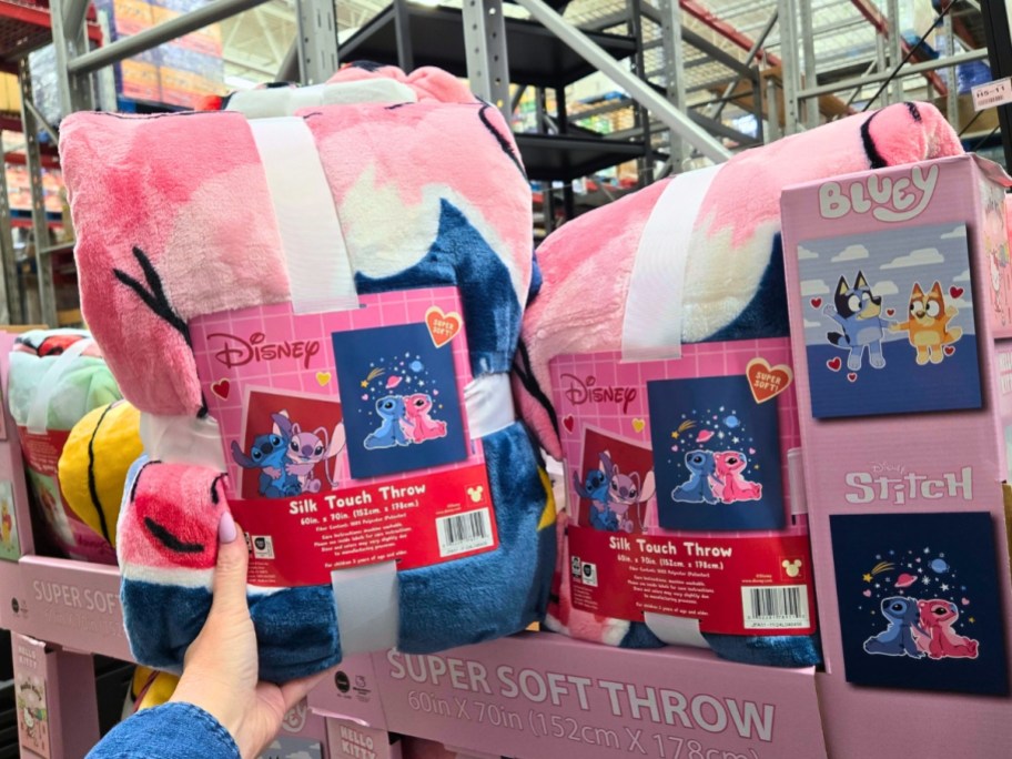 woman's hand holding up a Disney Stitch Valentine's themed plush throw blanket with a large bin of more character Valentine's Throw Blankets behind it