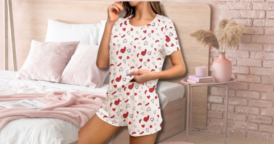 a woman wearing a pair of pajama shorts and a top in cream with red hearts, a bed with pink and white bedding is behind her