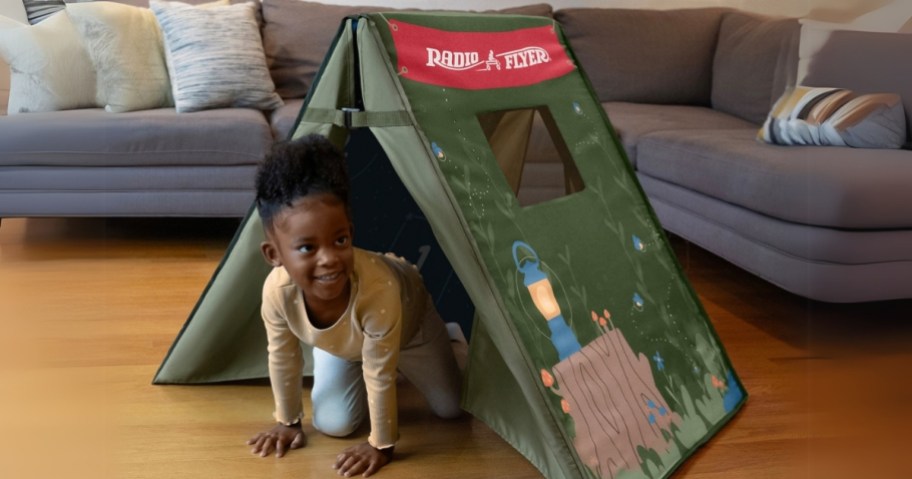 little girl crawling out of a green Radio Flyer play tent that's set up in a living room