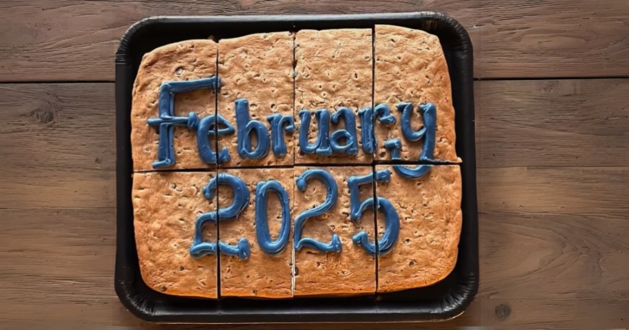 a cookie cake in a black tray on a woodtop table, the cake has blue icing with the words "February 2025" written on it