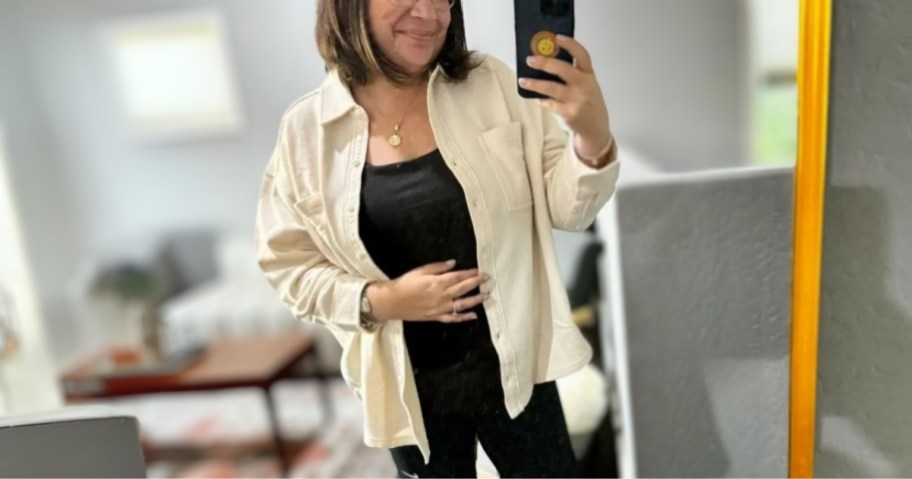 woman taking a selfie in a mirror wearing a cream color button up shirt with a black shirt and leggings