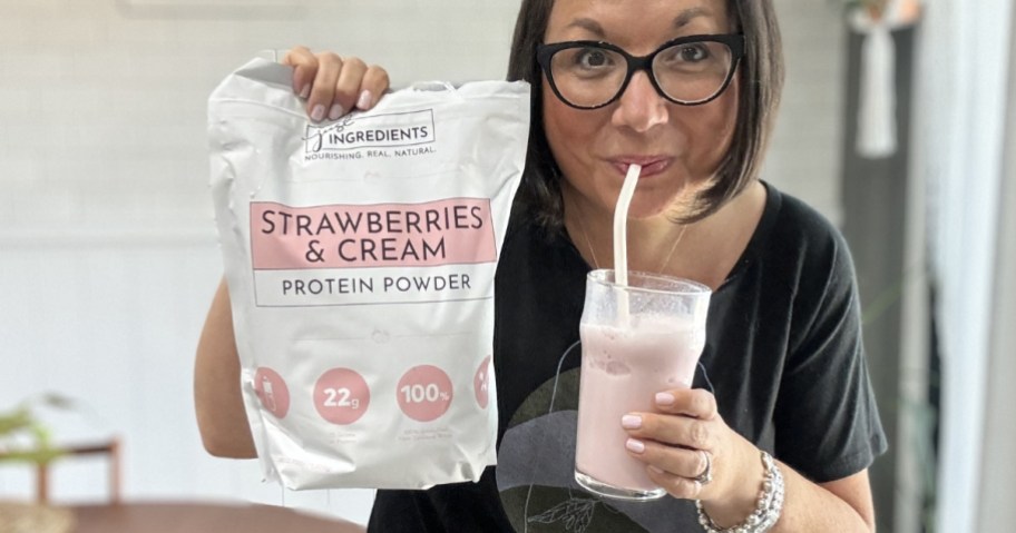 woman holding a bog of Strawberries & Cream protein powder <a href='https://tootightdu.com/blog/how-to-store-your-wig-while-traveling-the-ultimate-guide' target='_blank' rel='follow'>while</a> sipping on a straw from a glass with the protein powder” width=”912″ height=”479″></p>
<div class=