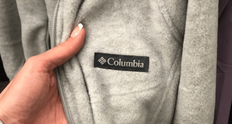 hand holding up a part of grey and white heather Columbia hoodie to show the Columbia logo