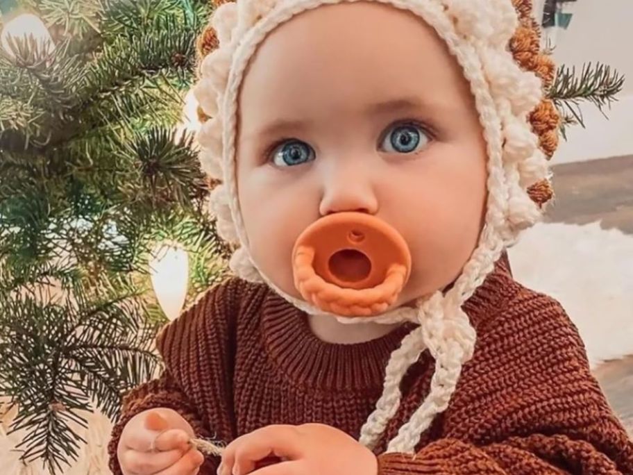 A baby sitting in front of a tree with a pacifier in her mouth 