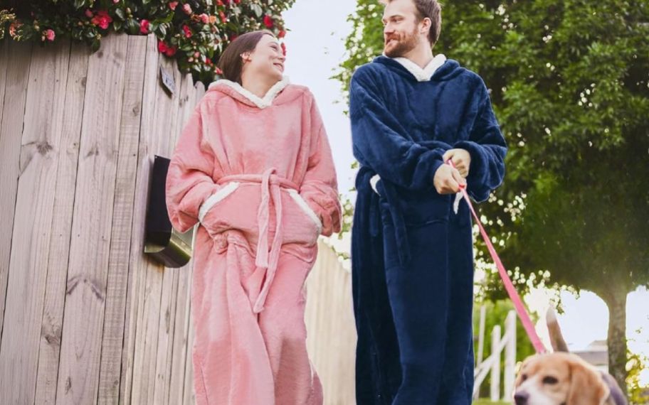 woman wearing pink wearable blanket and man wearing blue wearable blanket walking a dog