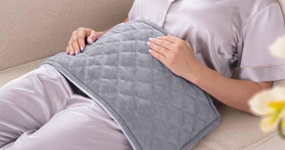 woman wearing button down light purple pajamas sitting on a tan couch with a grey quilted heating pad over her stomach