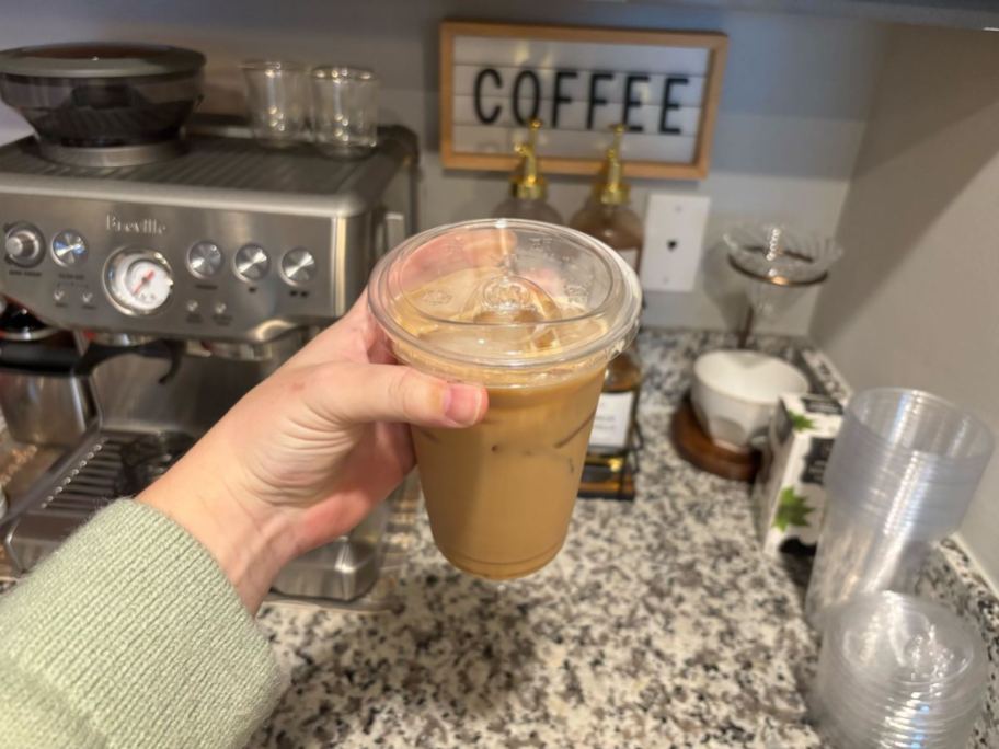 hand holding plastic cup with iced coffee in front of coffee station